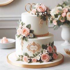 a three tiered wedding cake with pink flowers and gold rings on top, sitting on a table