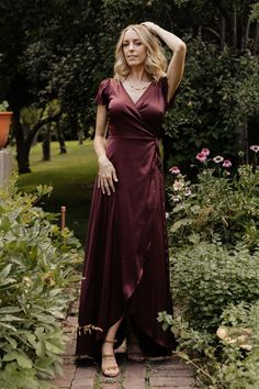 a woman in a long maroon dress standing on a brick path surrounded by greenery