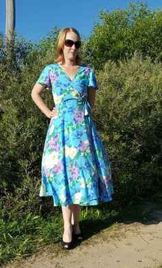 a woman standing in front of bushes wearing a blue dress with floral print on it