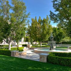 a fountain surrounded by hedges and trees in the middle of a lawn with benches on each side