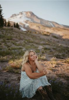 a woman sitting on the ground in front of a mountain with her hand under her chin