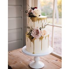 a three tiered cake with flowers and dripping glazes on the top, sitting on a wooden table in front of a window