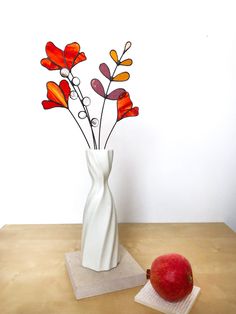 a white vase filled with red flowers next to an apple on a wooden counter top