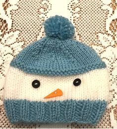 a blue and white knitted hat with a snowman face on the front, sitting on a lace doily