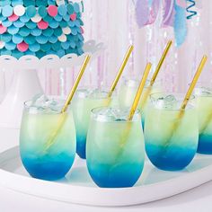 blue and green drinks with gold straws on a white tray in front of a cake