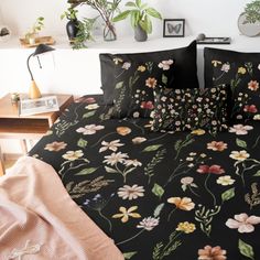 a bed covered in black floral sheets and pillows with plants on the wall behind it