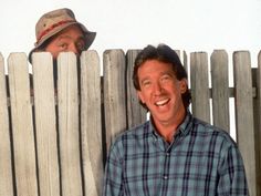 two men standing next to each other in front of a wooden fence with one smiling