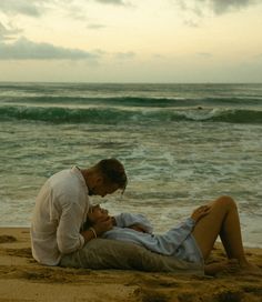 a man and woman are sitting on the sand by the ocean with their heads touching each other