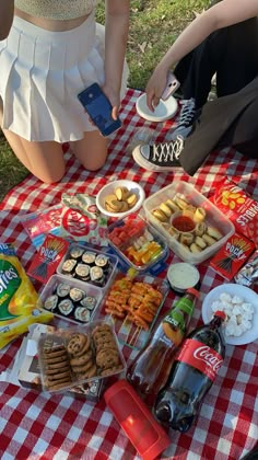 two people sitting at a picnic table with food and drinks on it, one person is holding a cell phone