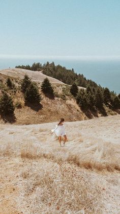 a woman walking across a dry grass covered hillside next to the ocean with trees in the background