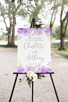 a welcome sign with purple flowers and greenery is displayed on a tripod stand