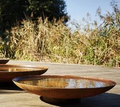 two brown bowls sitting on top of a wooden table next to tall grass and trees