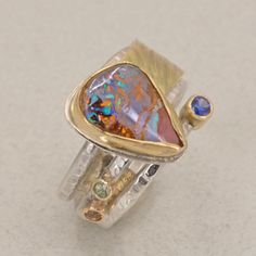 Boulder Opal set 18K Gold surrounded by textured Gold and Sterling with handmade triple silver band.  The ring in the first image and amethyst stone rings have both sold.  This listing is for the ring with the emerald stone or the sapphire stone.  This unique creation consists of a boulder opal set in a tall 18K gold bezel setting backed in sterling silver.  The opal is accented with a stone set off to the side set in 14K gold and a triple silver band which is handmade, formed, and textured.  Ad Unique Multi-stone Amethyst Ring For Anniversary, Unique Amethyst Multi-stone Rings, Fusion Style Opal Ring For Anniversary, Unique Multi-stone Gold Amethyst Ring, Unique Gold Multi-stone Amethyst Ring, Boulder Opal Ring, Oxidized Ring, Yellow Gold Setting, Emerald Stone