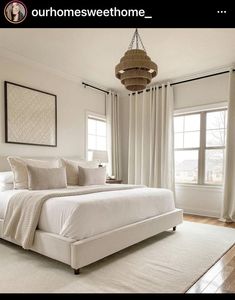 a white bed sitting in a bedroom next to two windows and a chandelier