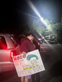 a woman holding a sign in the middle of a parking lot at night with cars behind her