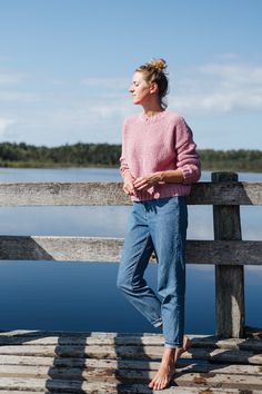 a woman is standing on a dock by the water wearing jeans and a pink sweater