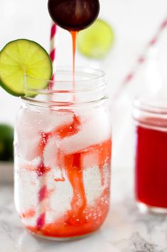 a mason jar filled with liquid, ice and lime wedges next to a red drink