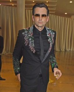 a man in a black suit and sunglasses holding a wine glass while standing on a wooden floor