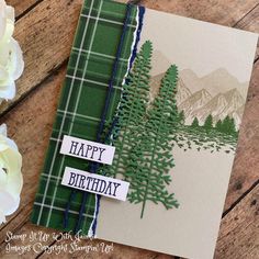 a happy birthday card with trees and mountains in the background on a wooden table next to flowers