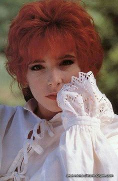 a woman with red hair wearing a white blouse and holding her hand to her face