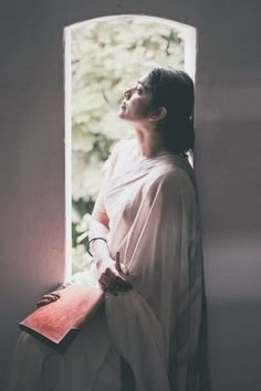 a woman standing in front of a window with her eyes closed and holding a book