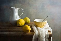 two lemons and a bowl on a table with a white pitcher next to it