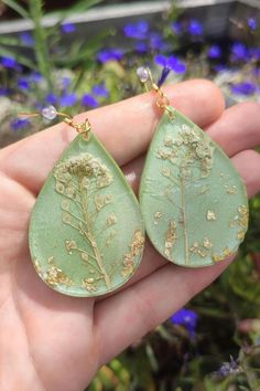 a hand holding a pair of green earrings with gold leaf designs on it and blue flowers in the background