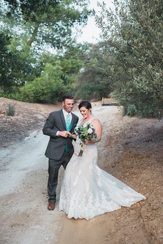 a bride and groom walking down a dirt path in front of an olive tree at their wedding