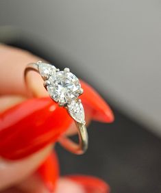 a woman's hand holding an engagement ring with three diamonds on it and red nail polish