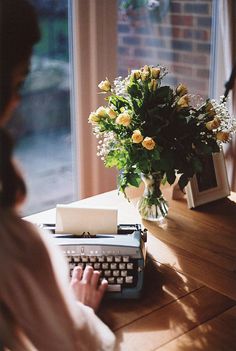 a woman typing on an old - fashioned typewriter next to a vase with flowers