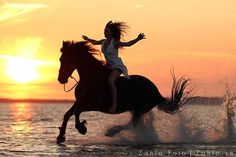 a woman riding on the back of a black horse in the ocean at sunset or dawn