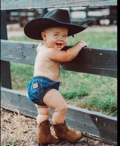 a baby boy wearing a cowboy hat leaning against a fence