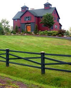 a large red house sitting on the side of a lush green field next to a wooden fence