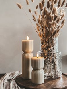 three white candles sitting on top of a wooden table next to a vase filled with dry grass