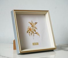 a white and gold framed flower in a wooden frame on a marble countertop,