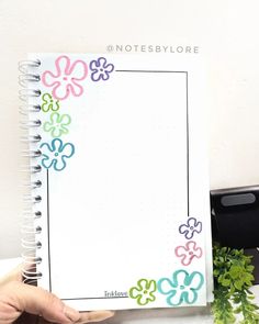 a person holding up a notebook with flowers on the cover and writing in it, next to a potted plant