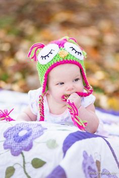 a baby wearing a knitted owl hat while laying on a blanket in the woods