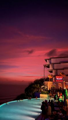 people are sitting on the edge of a swimming pool at night with colorful clouds in the background