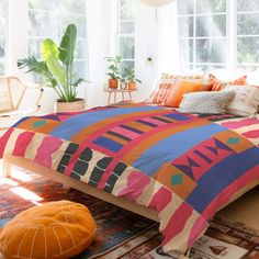 a bed with colorful comforter and pillows in a room next to a plant on the floor