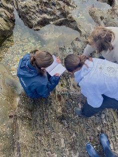 two girls are looking at something in the water on some rocks and sand with their hands together