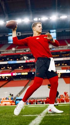 a man in red shirt and black pants holding a football on field with stadium lights behind him