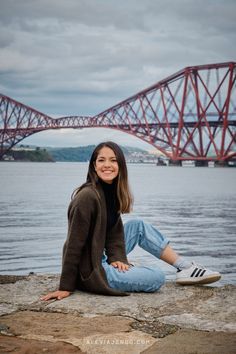 a woman sitting on top of a rock next to the water with a bridge in the background