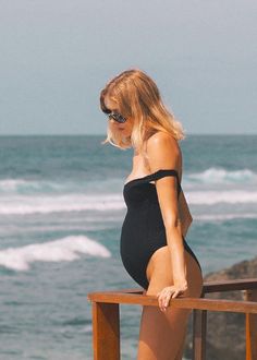 a woman in a black swimsuit standing on a wooden railing next to the ocean