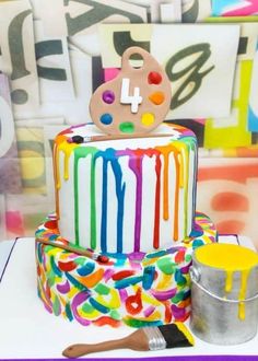 a multi - colored birthday cake with icing dripping from it and a paintbrush next to it