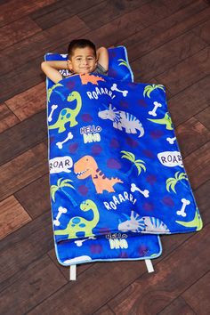 a young boy laying on top of a blue dinosaur print sleeping bag in the middle of a wooden floor