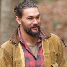 a man with a beard wearing a brown jacket and plaid shirt standing in the woods