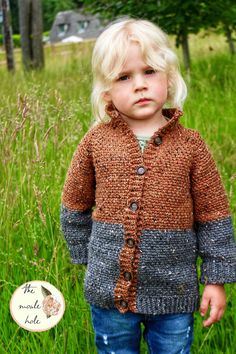 a little boy standing in the grass wearing a brown and gray sweater with buttons on it