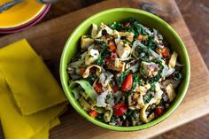 a green bowl filled with pasta and vegetables on top of a wooden cutting board next to a yellow napkin