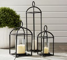 two black metal lanterns with candles in them sitting on the ground next to a potted plant