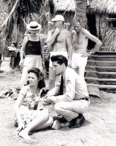 a group of people standing and sitting on the ground in front of some huts with palm trees